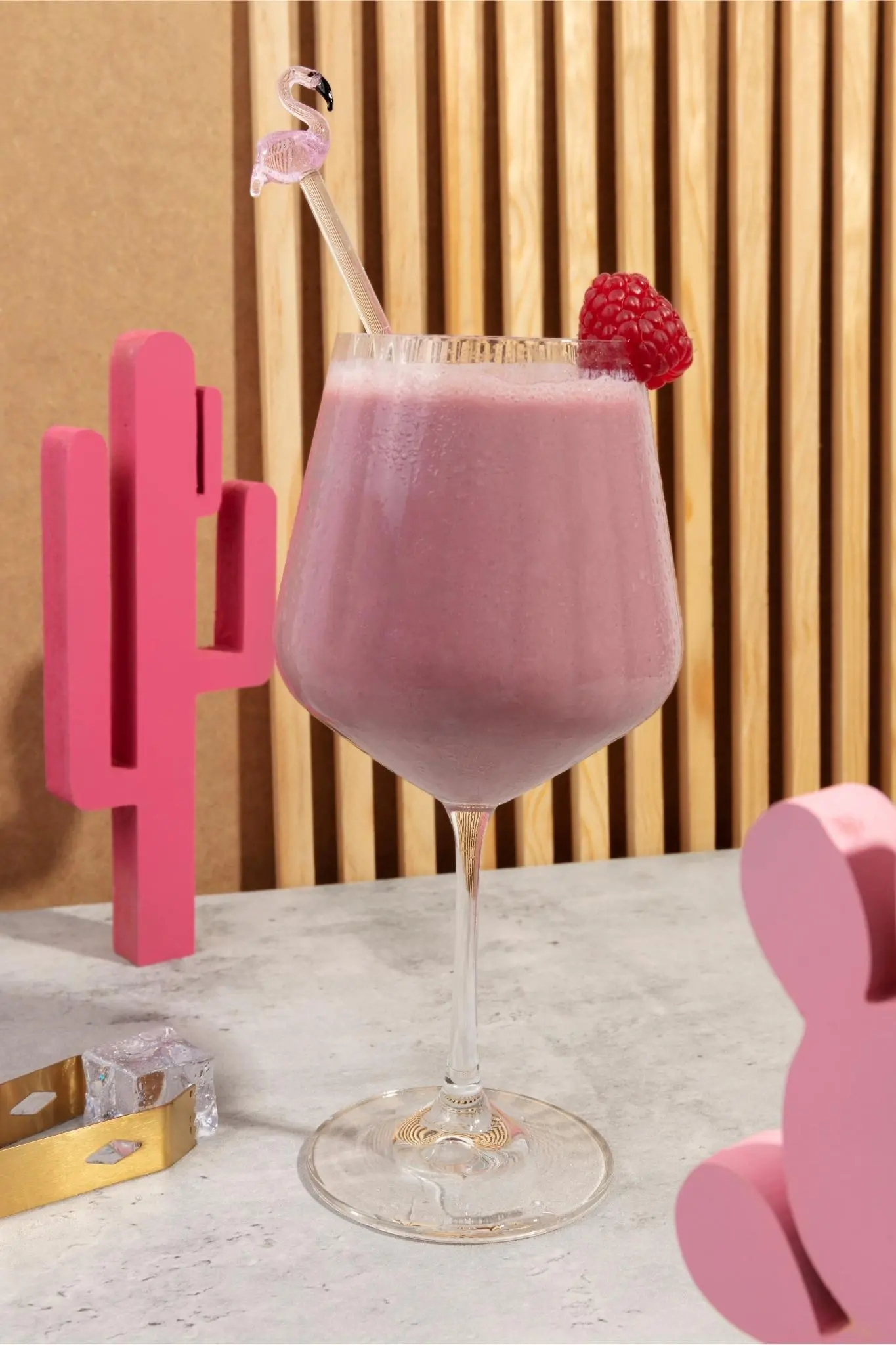 A vibrant pink drink served in a clear wine glass, garnished with a raspberry and a flamingo-themed stirrer, with modern wooden decor in the background