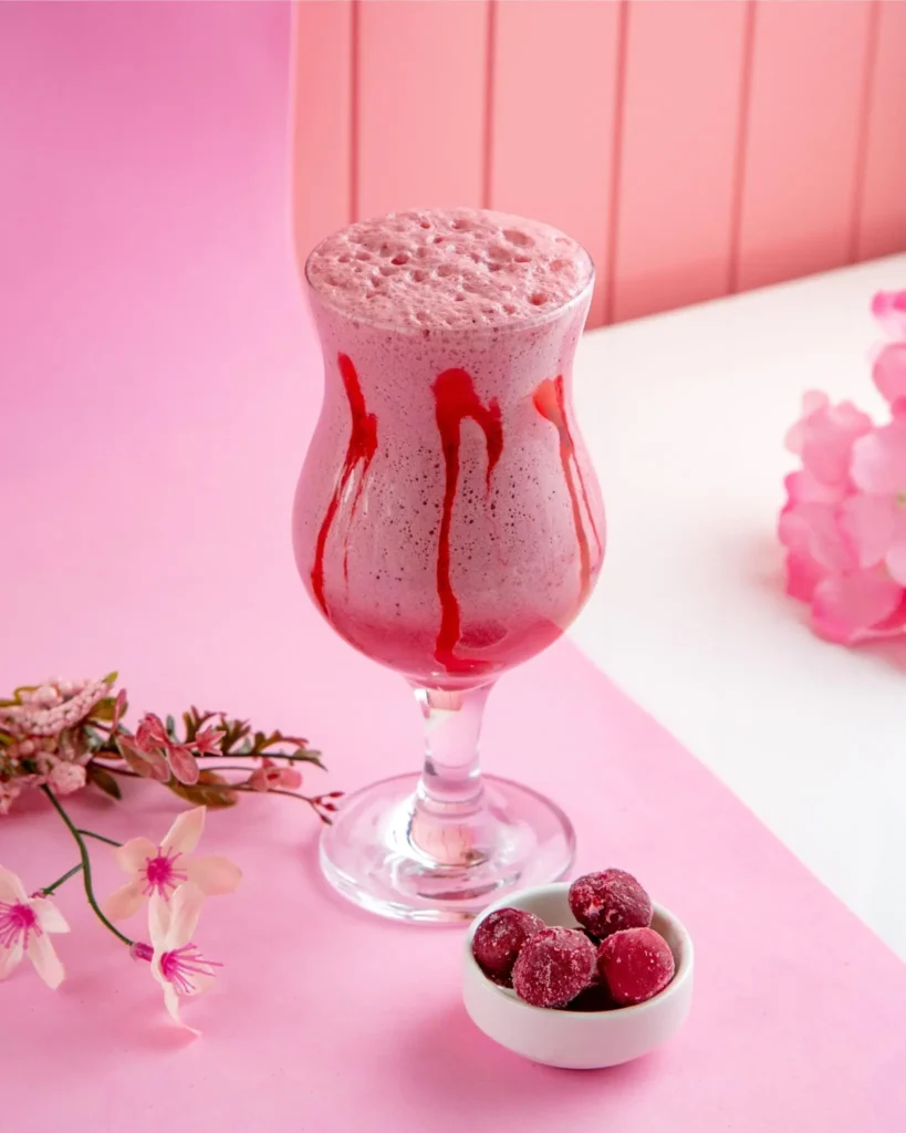 A pink smoothie in a decorative glass with red syrup drizzles, garnished with frozen cherries and surrounded by pink flowers on a pastel pink background.