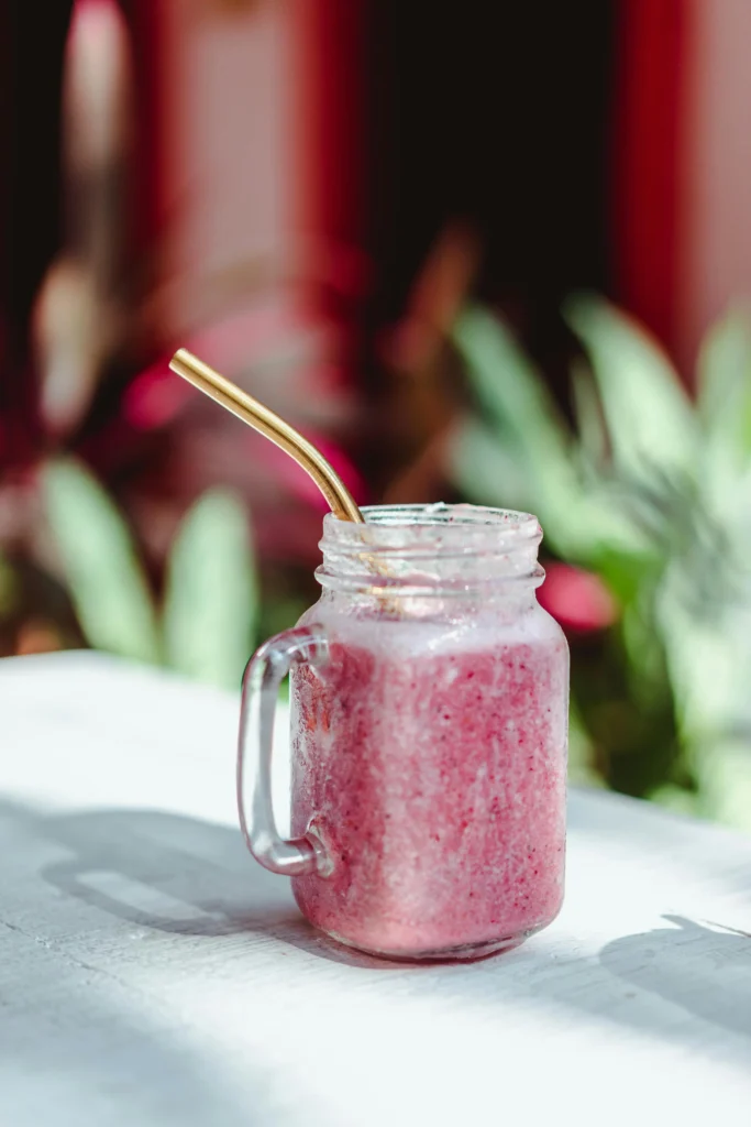 A refreshing pink drink served in a glass mason jar with a metal straw, sitting on a white table under natural sunlight, surrounded by blurred greenery in the background.