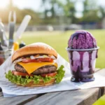 A grilled burger and a grape snow cone on a picnic table with a summer backdrop.