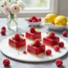 Four rectangular raspberry lemon mousse cakes on a white surface, garnished with fresh raspberries, white chocolate, and herbs.