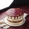 Multi-layered cake with deep maroon and off-white frosting, adorned with gold leaf, on a maroon surface.